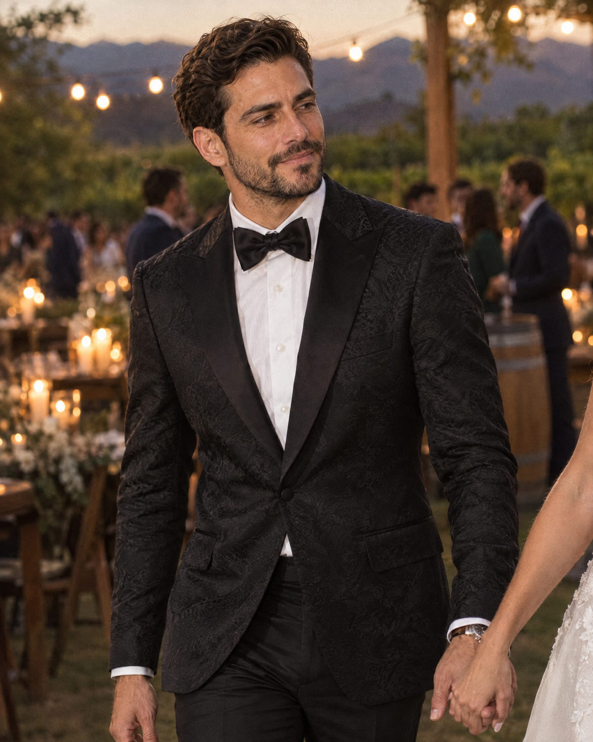 Novio con esmoquin Imperiale Negro de jacquard tomado de la mano de la novia en una viña